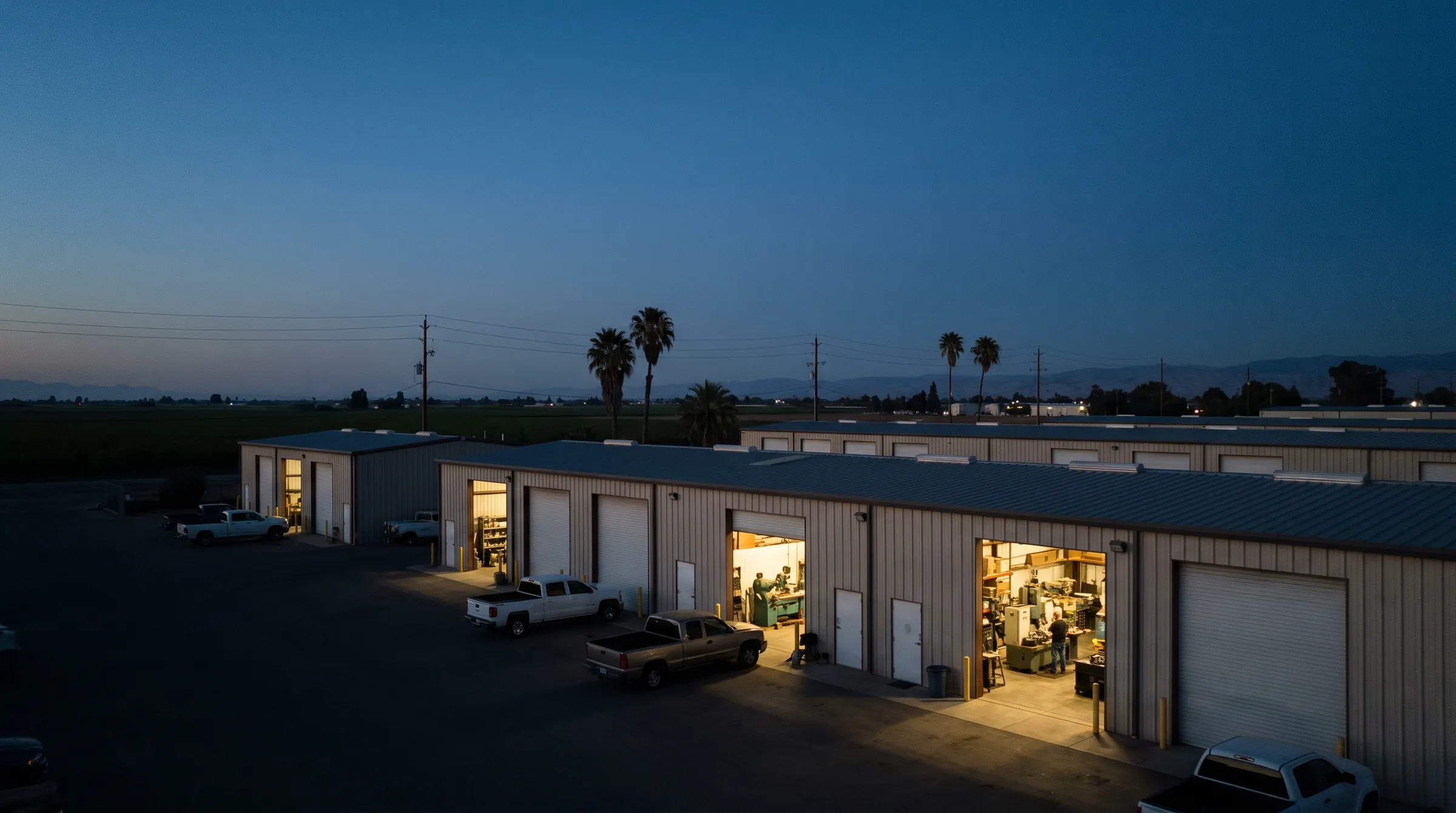 Rows of small metal shop buildings in a Central Valley industrial park at dusk, lights on in the open bays.